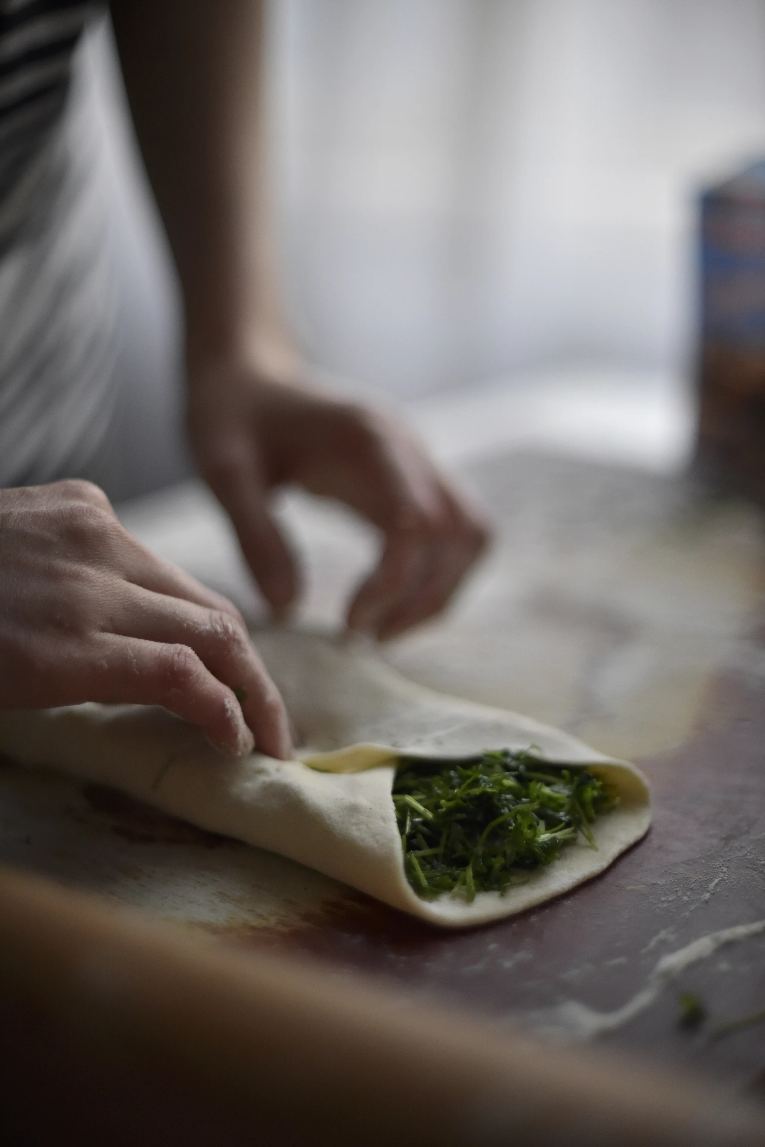 A hand folding dough around a green filling on a worktable.